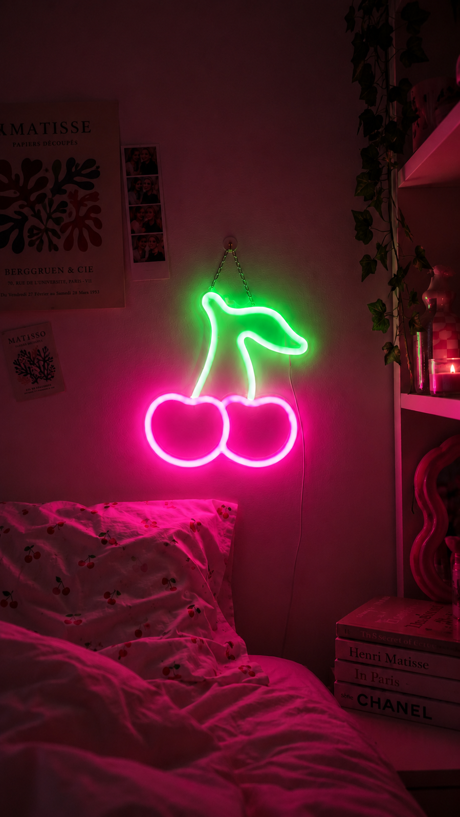 A neon wall light shaped like two cherries, glowing in bright pink and green, mounted on a bedroom wall above a bed.
